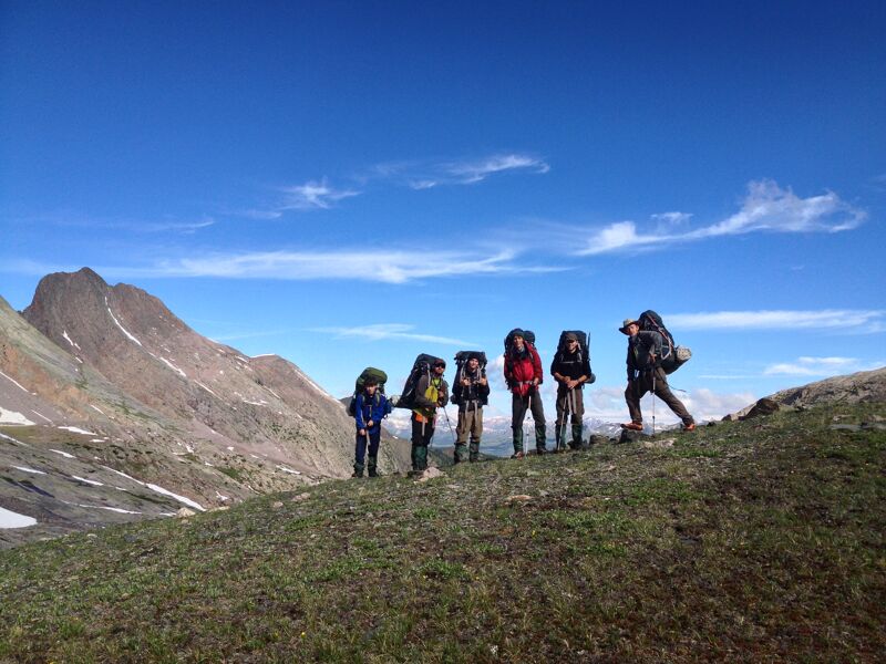 A group of seven people are hiking on a grassy hillside with mountains in the background. They are all wearing large backpacks, suggesting they are on a multi-day trip. The sky is blue with some scattered clouds. The landscape appears to be in a mountainous region, possibly during the summer months, given the green vegetation.